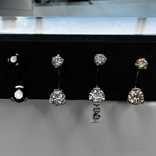 A set of three body jewelry bars with prong-set clear, champagne, and black colored gems, displayed on a black stand.